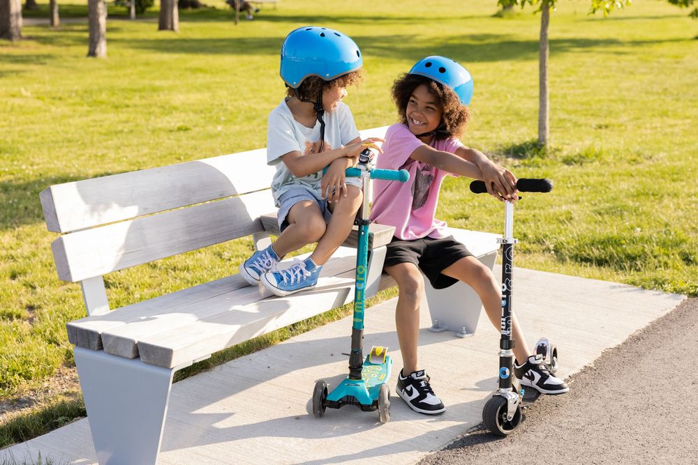 Two children wearing blue helmets sit on a park bench with teal kick scooters on a sunny day.