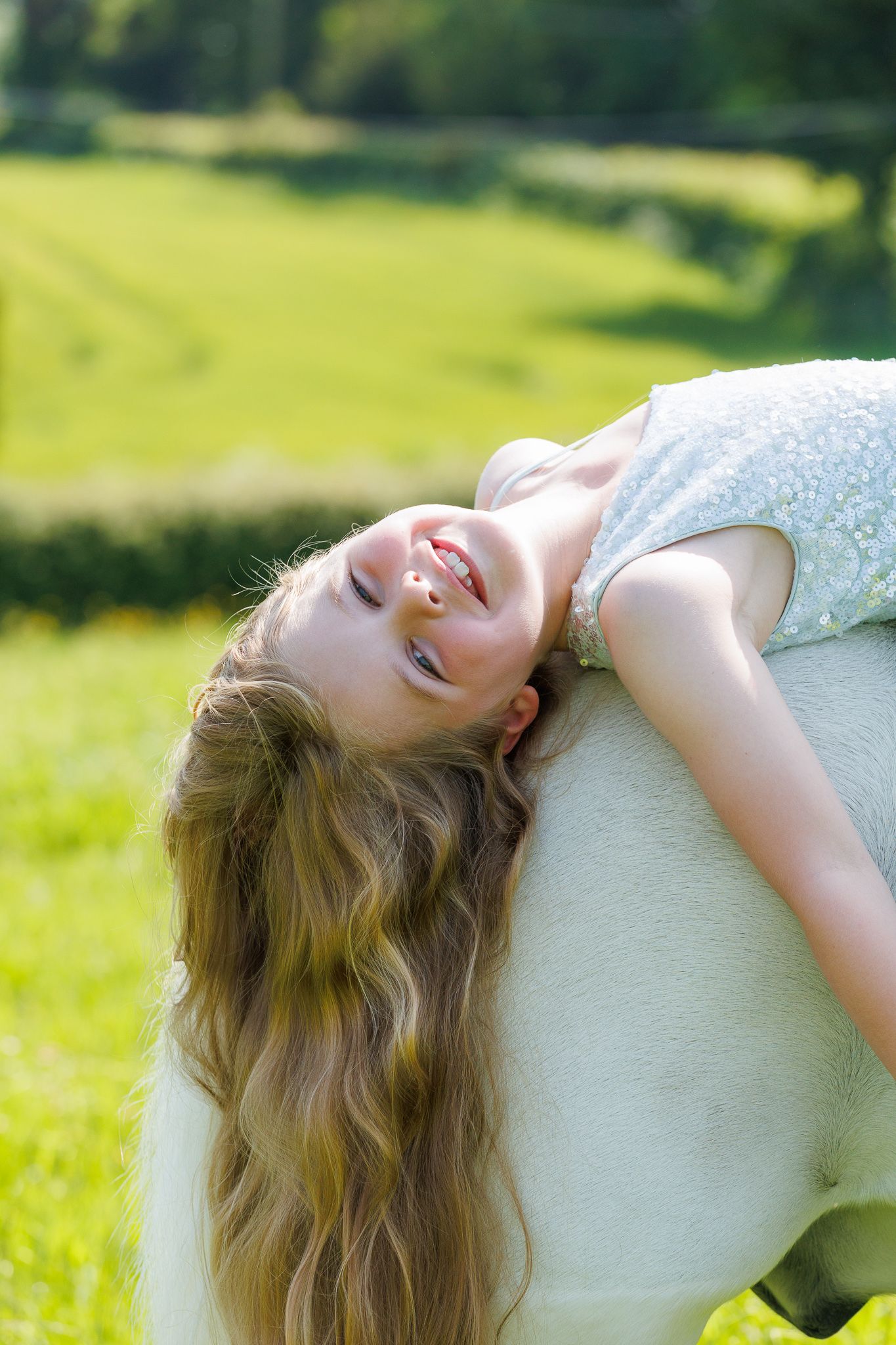 Young girl with long blonde hair laughing joyfully while leaning back outdoors in a sunny green meadow.