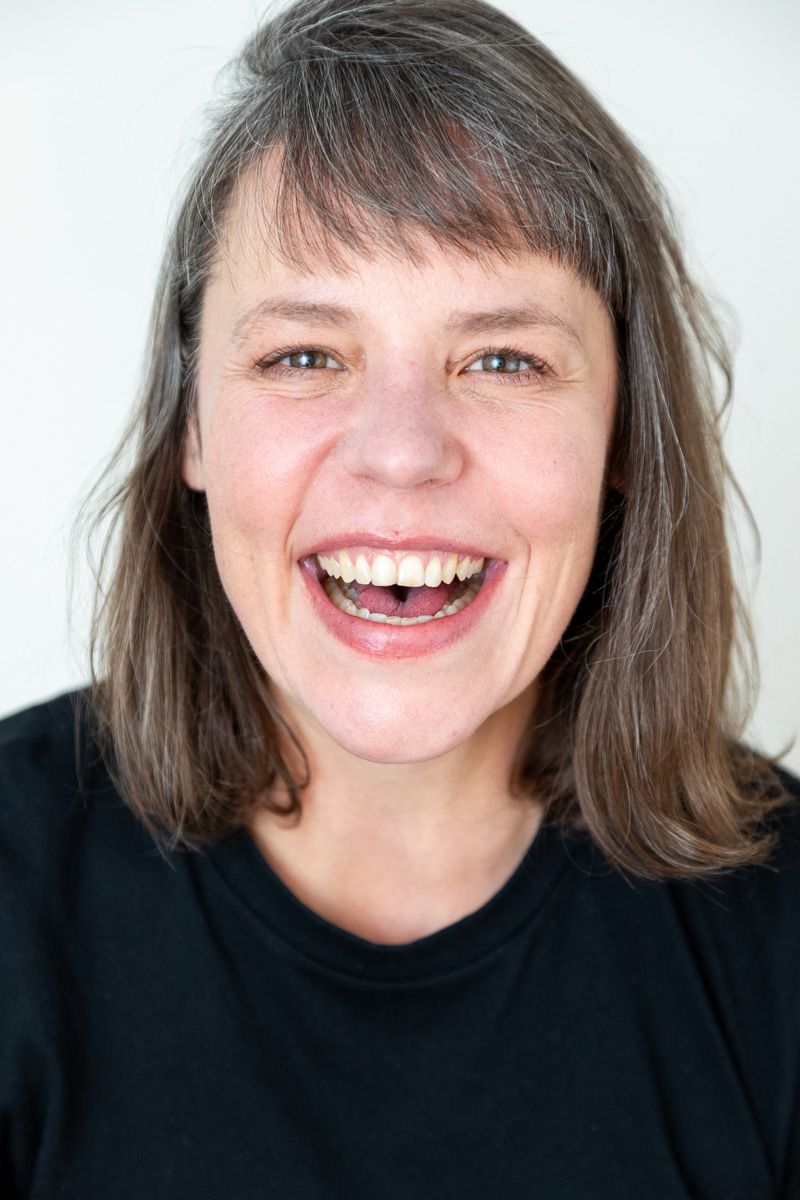 A middle-aged woman with brown hair laughing joyfully, wearing a black top against a white background.
