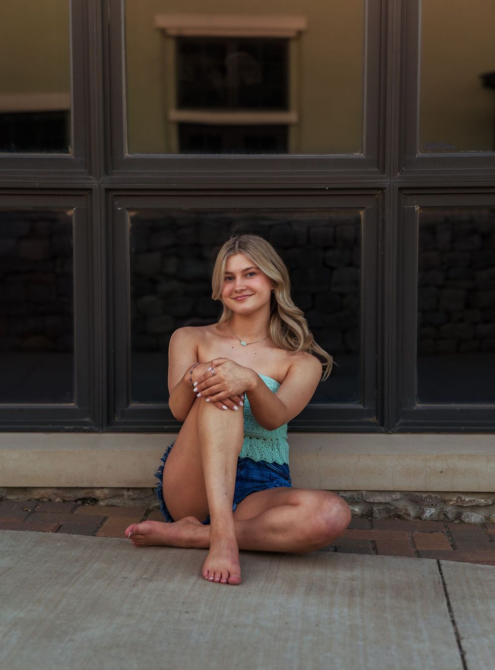 Young woman in teal top and denim shorts sitting barefoot on brick pavement in front of large windows.