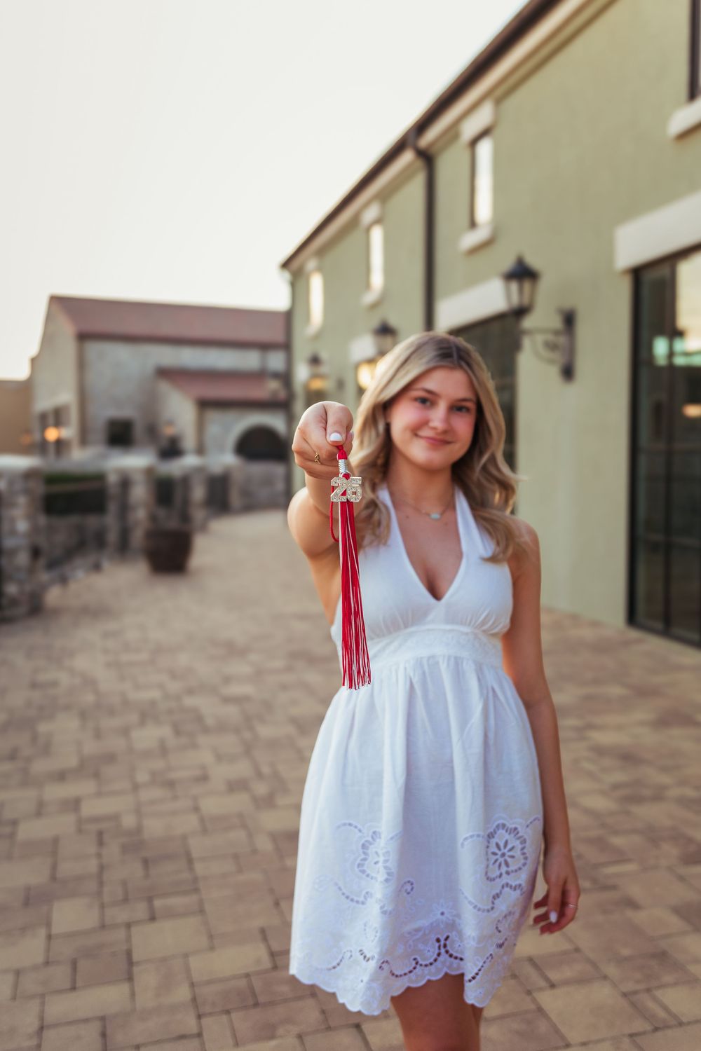 Young blonde woman in white halter dress holding red tassel keychain outdoors near elegant building.