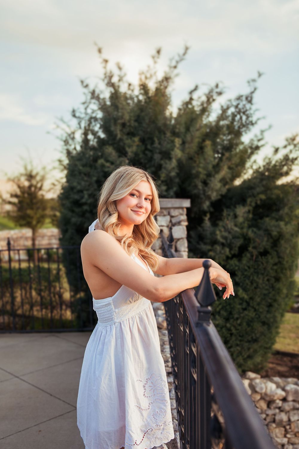 Young blonde woman in white sundress smiling on outdoor balcony with iron railing and green trees.
