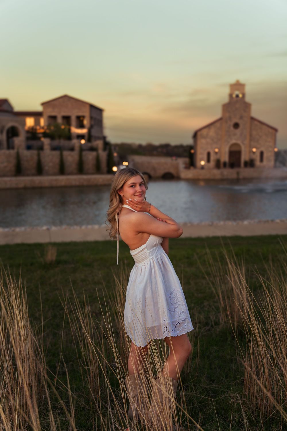 Young woman in white dress poses at sunset near a scenic pond with stone buildings in background.