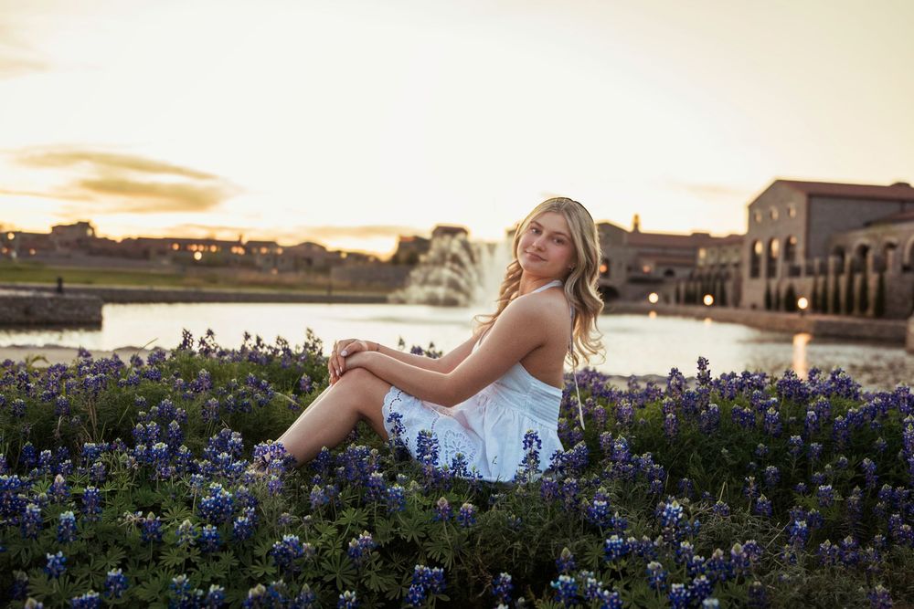 Young woman in white dress sitting among bluebonnets at sunset near a fountain.