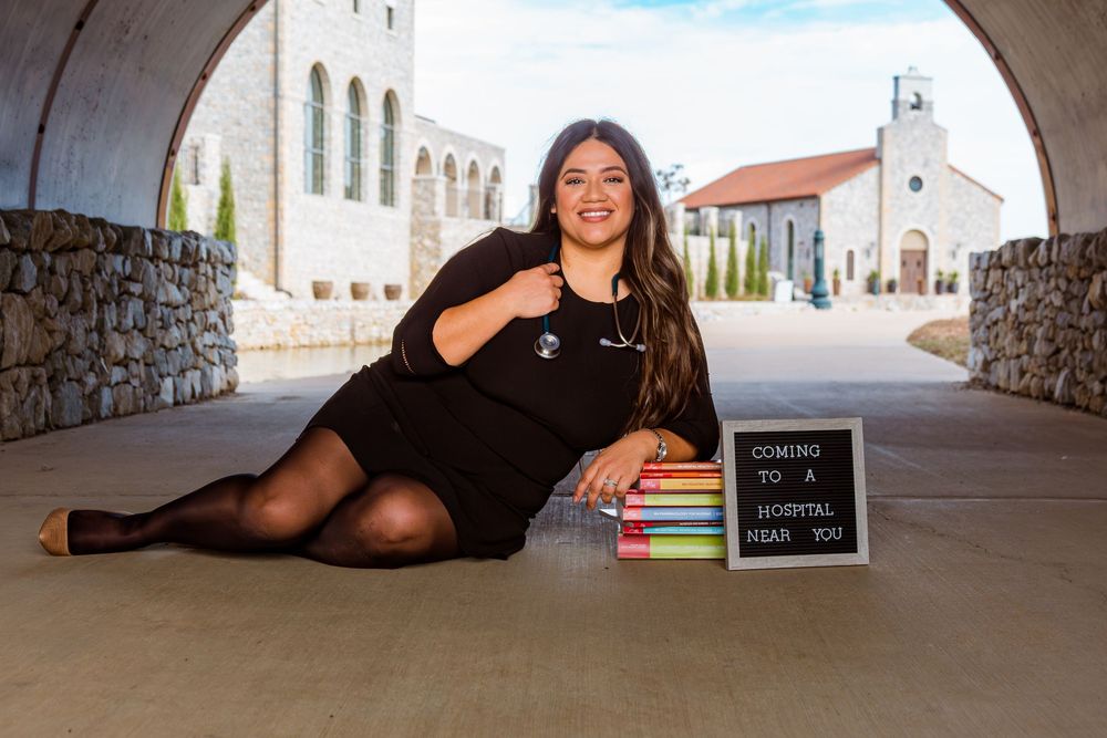 Woman in black dress poses with stethoscope and books beside a "Coming to a Hospital Near You" sign.