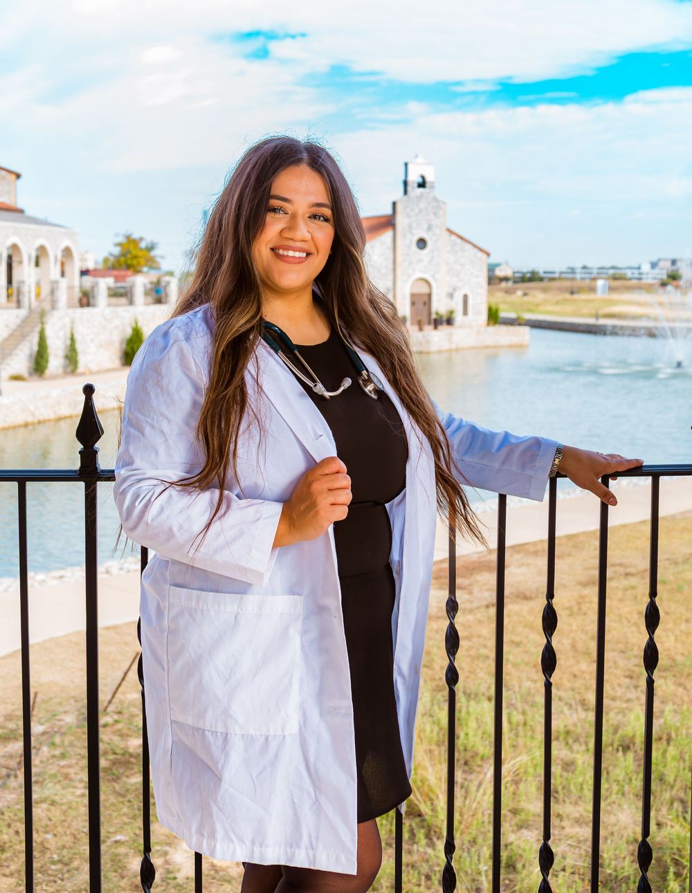 Young woman in white lab coat smiling outdoors near waterfront with historic building backdrop on sunny day.