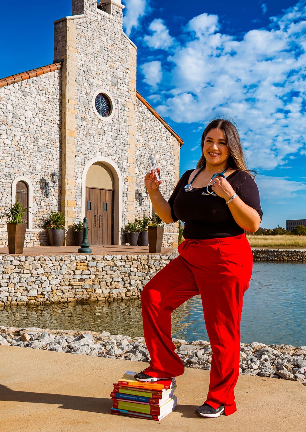 Woman in red scrubs holding stethoscope poses with medical books outside a stone church near a pond.