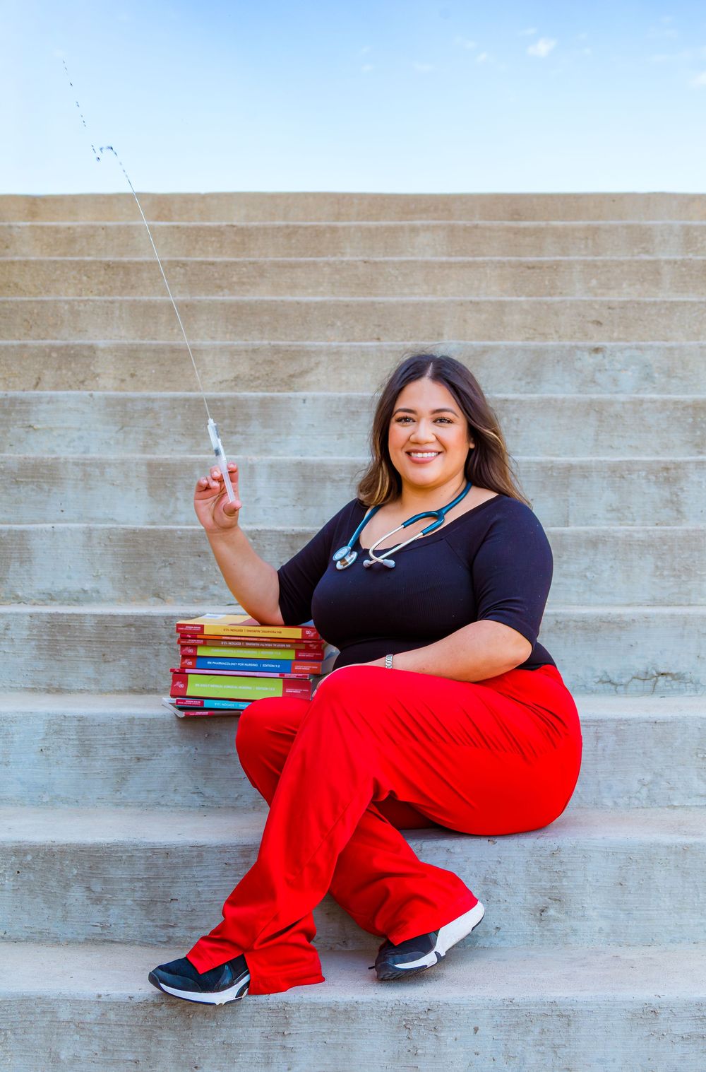 A smiling woman in red scrubs and stethoscope sits on steps holding a pen beside a stack of colorful textbooks.