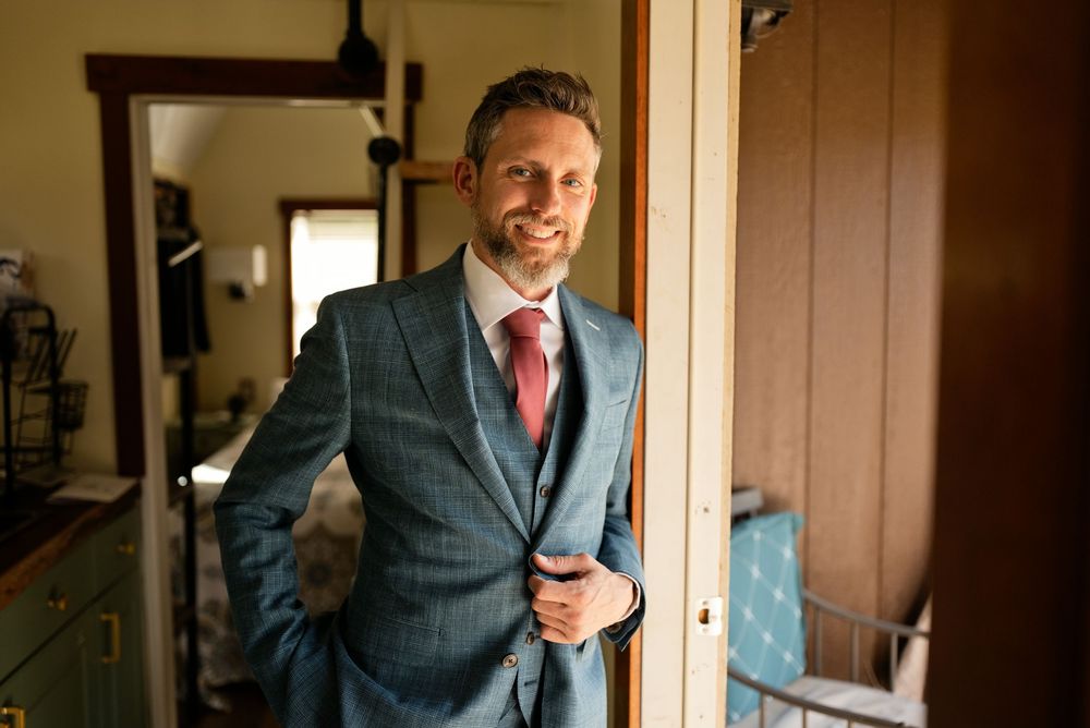 Smiling groom in a blue plaid three-piece suit with pink tie posing indoors on his wedding day.