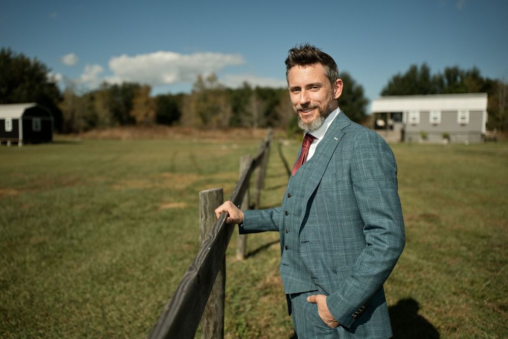 Elegant groom in blue plaid suit leaning on a rustic fence in an open countryside field.