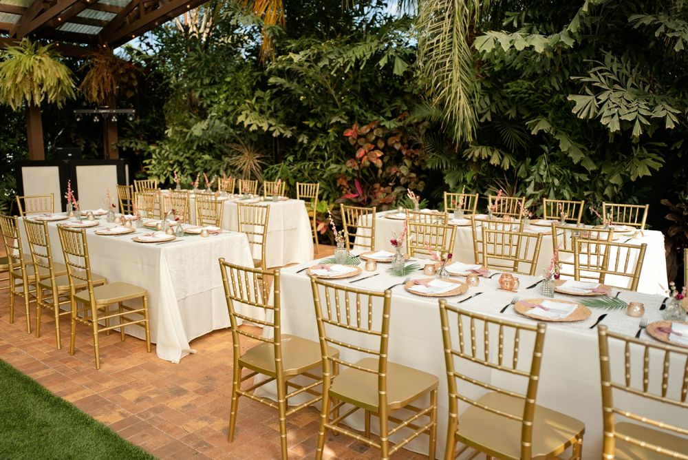 Elegant outdoor wedding reception with gold Chiavari chairs and white tablecloths surrounded by lush tropical greenery.