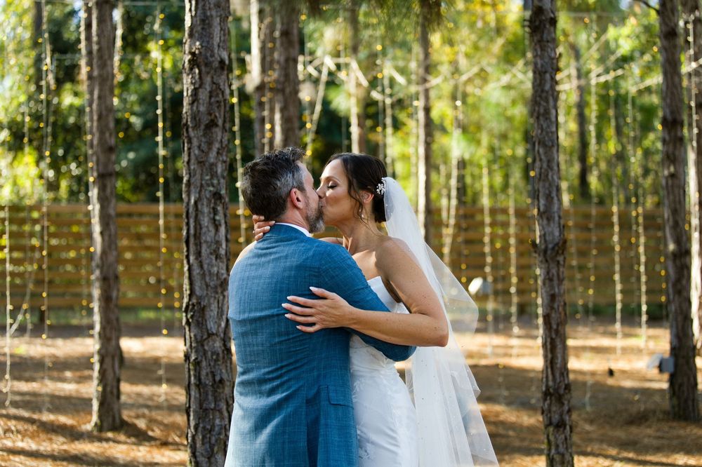 A bride and groom share a romantic kiss among tall pine trees decorated with hanging lights at their outdoor wedding.