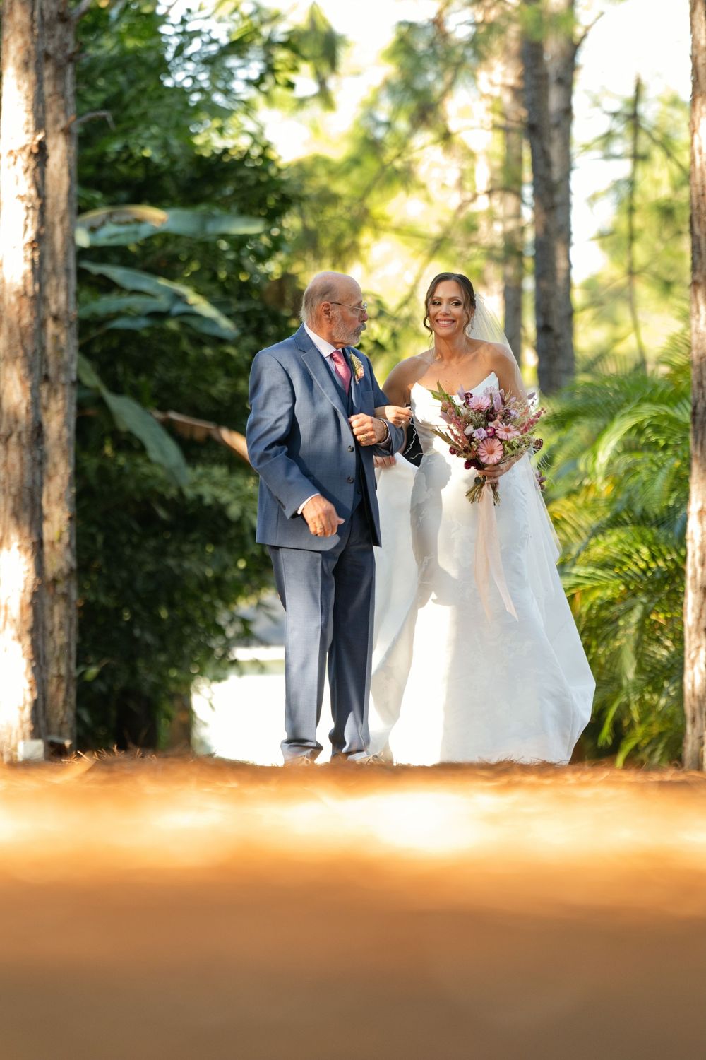 Bride in white gown walks with older man in blue suit through lush green tree-lined path holding pink bouquet.