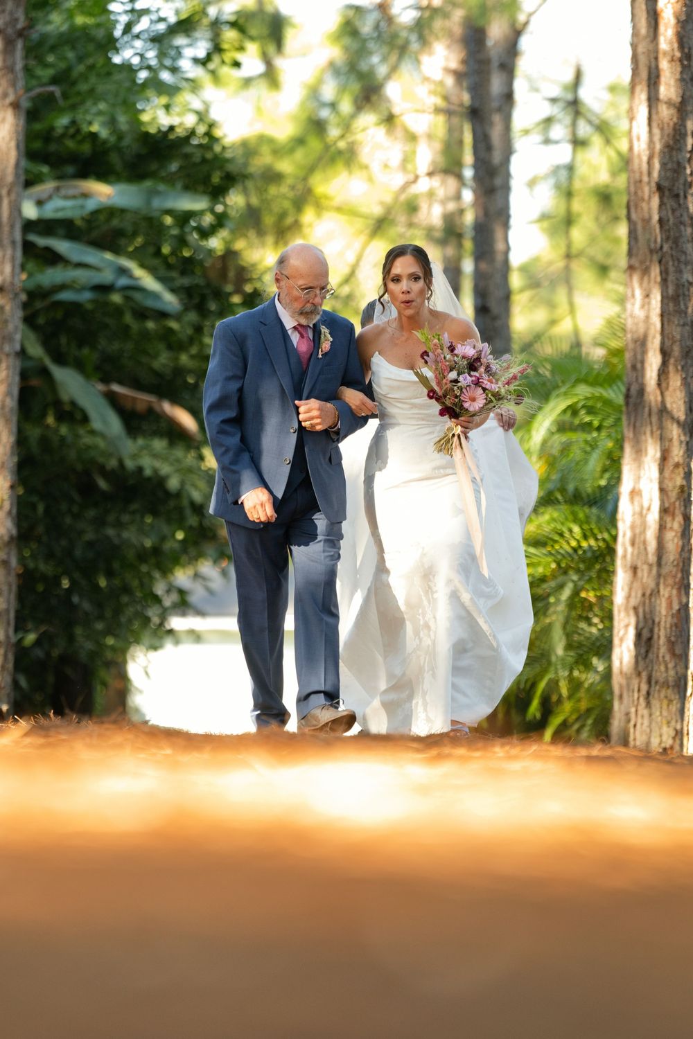 Bride in white gown holding bouquet walks arm-in-arm with older man in blue suit through sunlit tree-lined path.