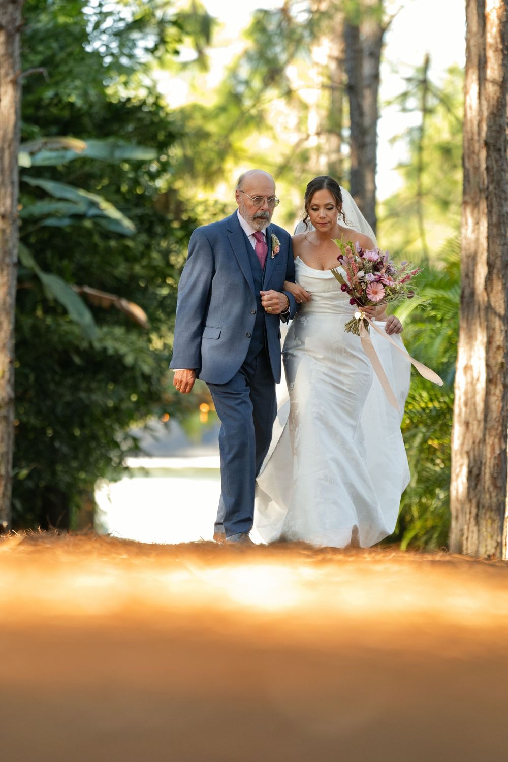 Bride in white gown walks with older man in gray suit through sunlit tree-lined path, holding pink bouquet.