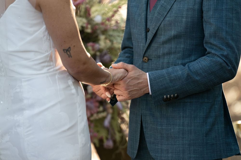 Bride and groom exchanging rings during wedding ceremony, bride showing butterfly tattoo on arm, groom in blue suit.