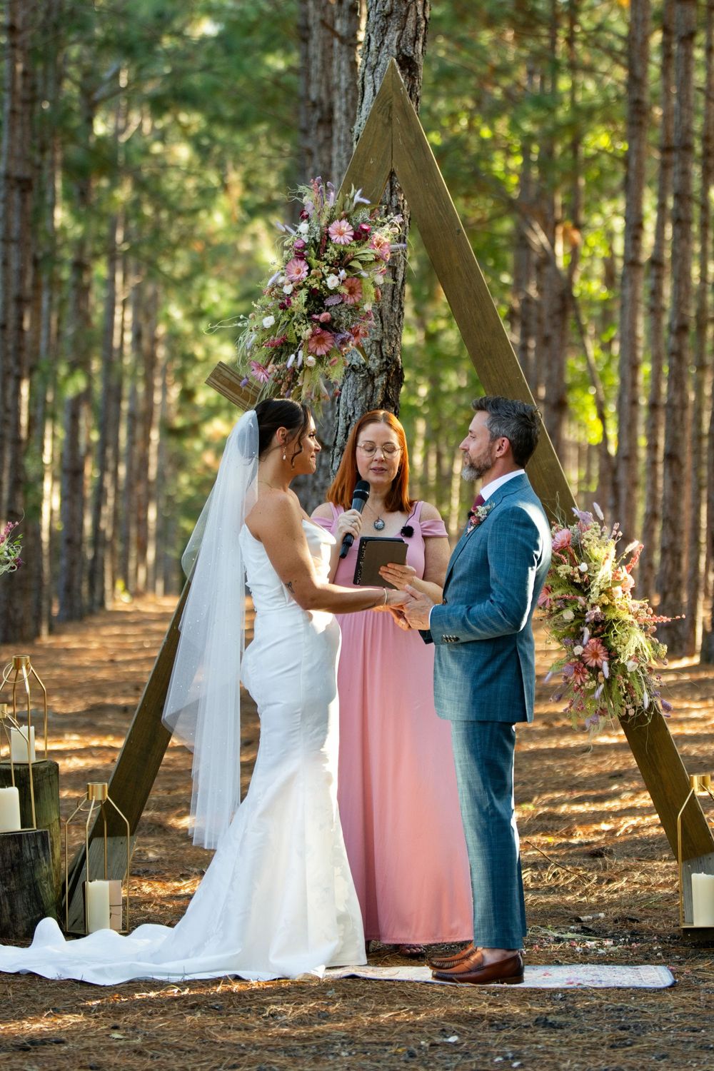 Couple exchanges vows at outdoor forest wedding ceremony under triangular arch with pink floral arrangements and officiant.