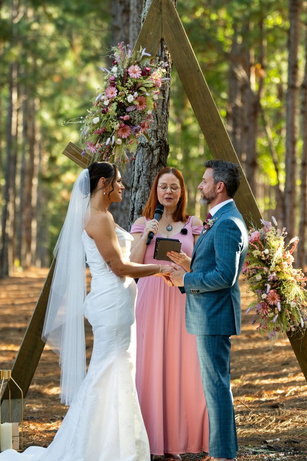 Bride and groom exchange vows outdoors in a forest wedding ceremony beneath a triangular wooden arch with pink floral decor.
