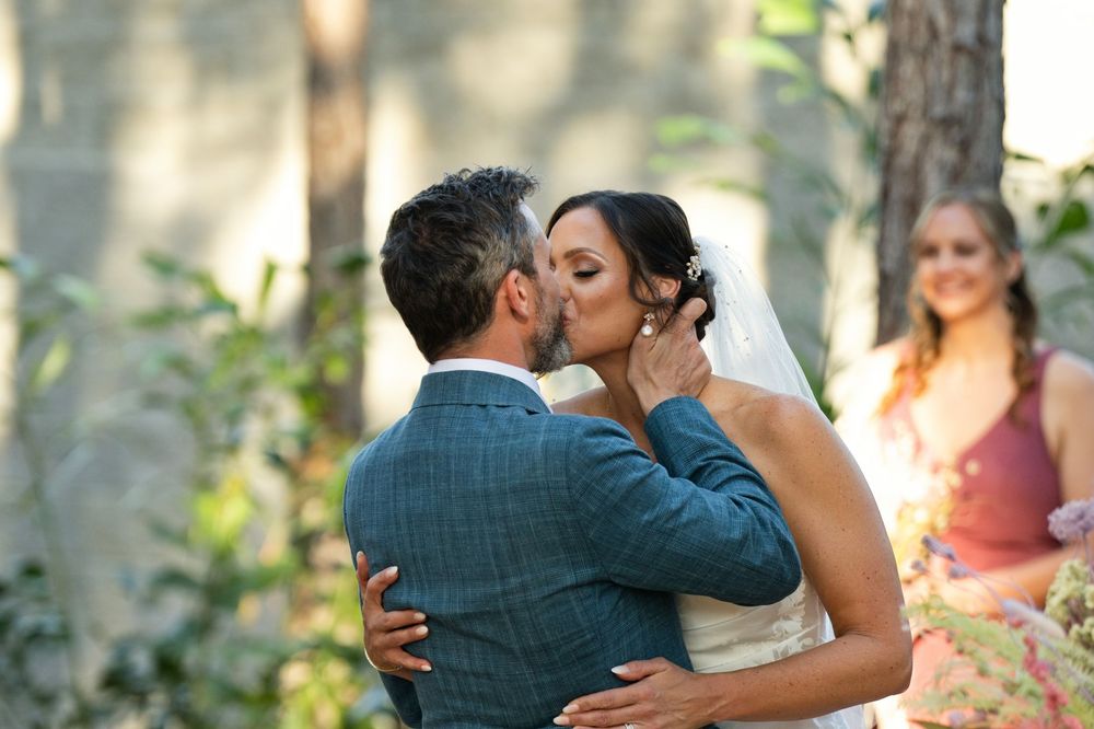 A bride and groom share their first kiss at an outdoor wedding ceremony surrounded by trees.