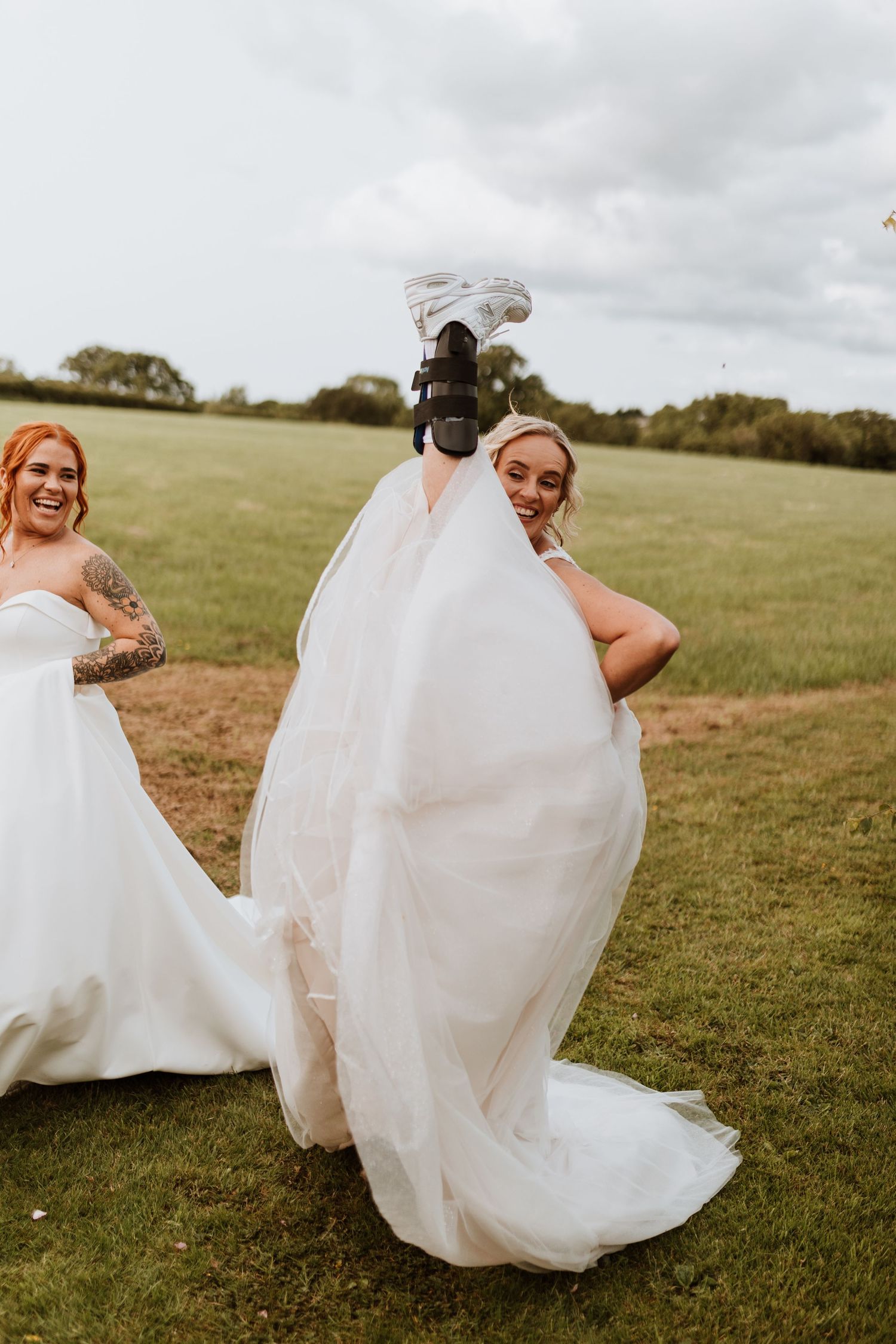 Two brides in white gowns laugh outdoors, one doing a high kick in dress to reveal black boots on a green field.