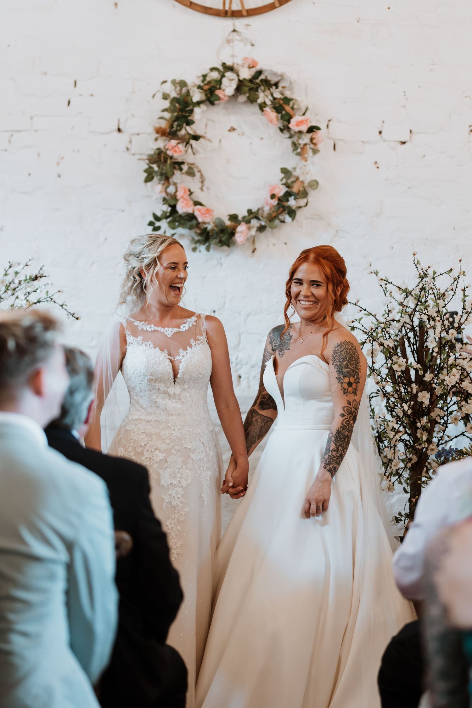 Two brides in white wedding gowns hold hands at the altar, smiling joyfully before a floral wreath backdrop.