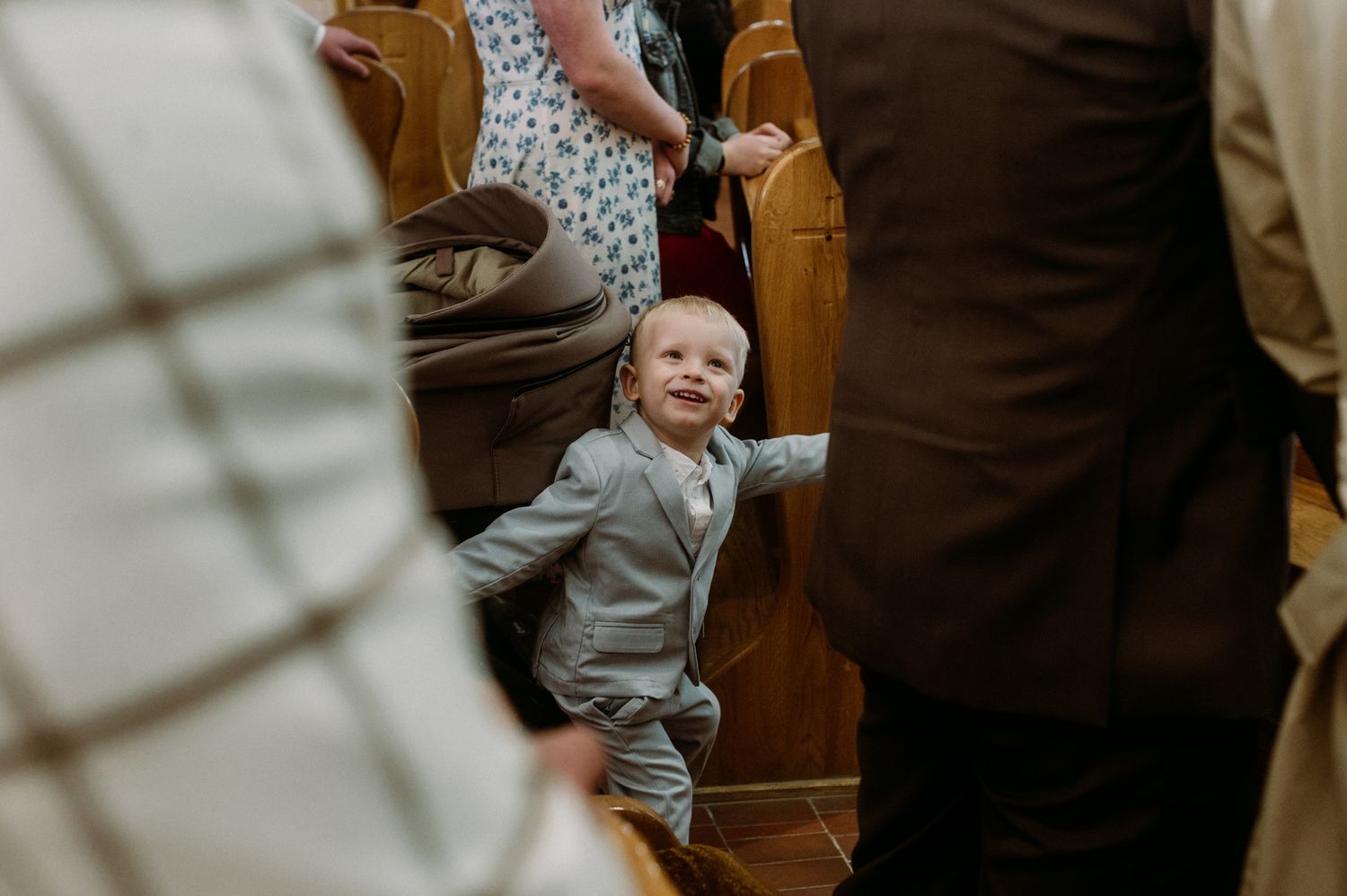 Adorable toddler boy in gray suit smiling at wedding ceremony between wooden church pews.