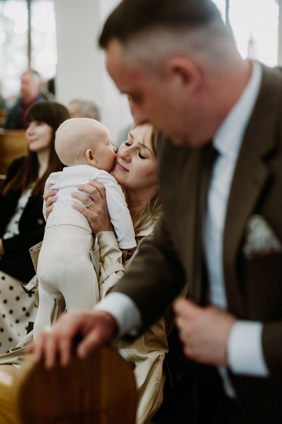 A mother tenderly kisses her baby during a baptism ceremony in a church, with a man in a suit visible in the foreground.
