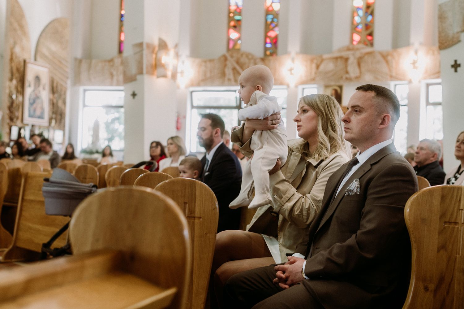Family attending a church ceremony, woman holding baby in wooden pew-filled sanctuary with stained glass windows.