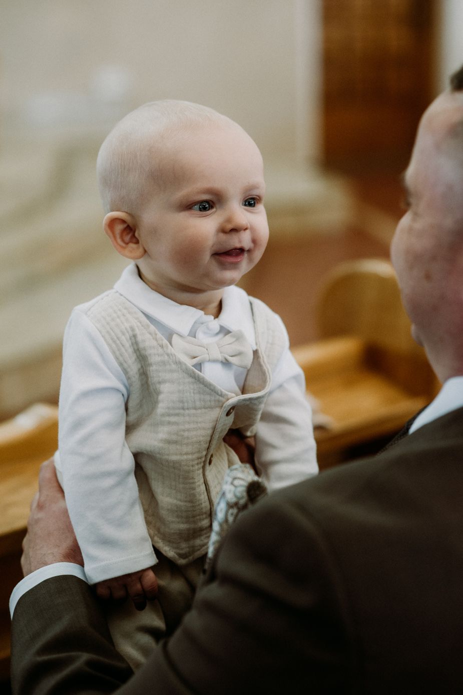 Adorable baby boy in a white shirt and bow tie vest smiles while being held at a formal event.