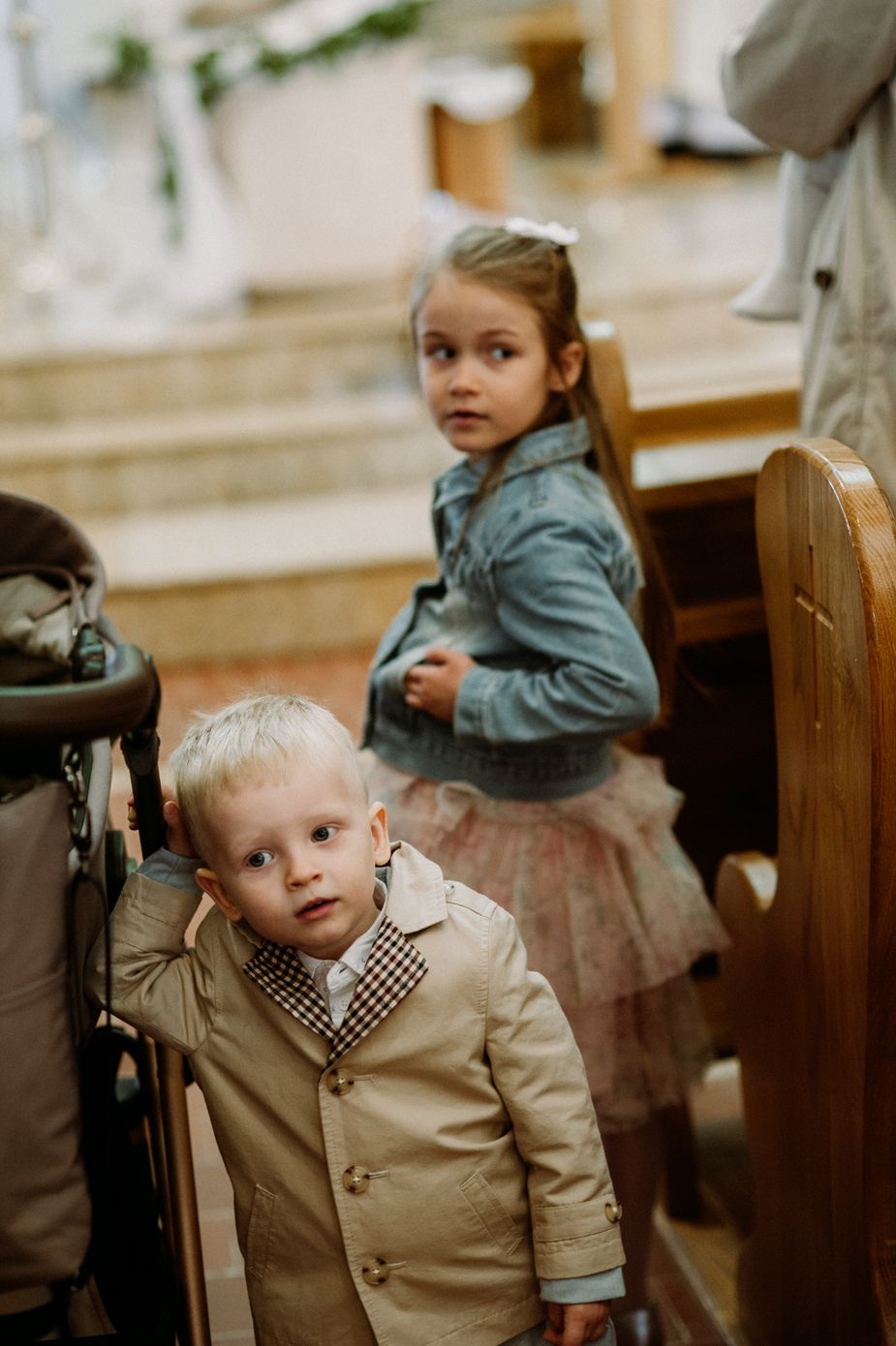 Two young children dressed in formal attire stand near wooden church pews, a boy in a tan coat and girl in a blue jacket.