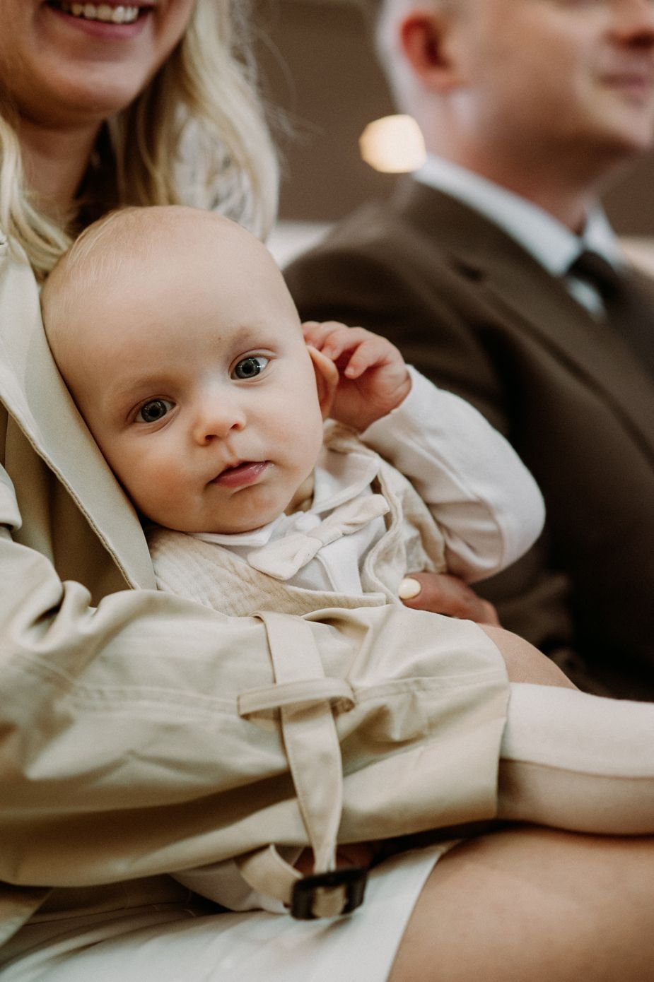 A cute baby in a white outfit rests in the arms of a woman wearing a beige trench coat at a formal event.