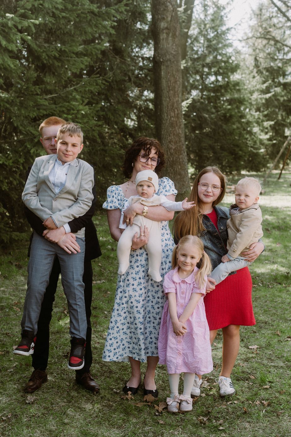 Family portrait with woman holding baby, teen girl with toddler, and boy carrying child, outdoors near pine trees.