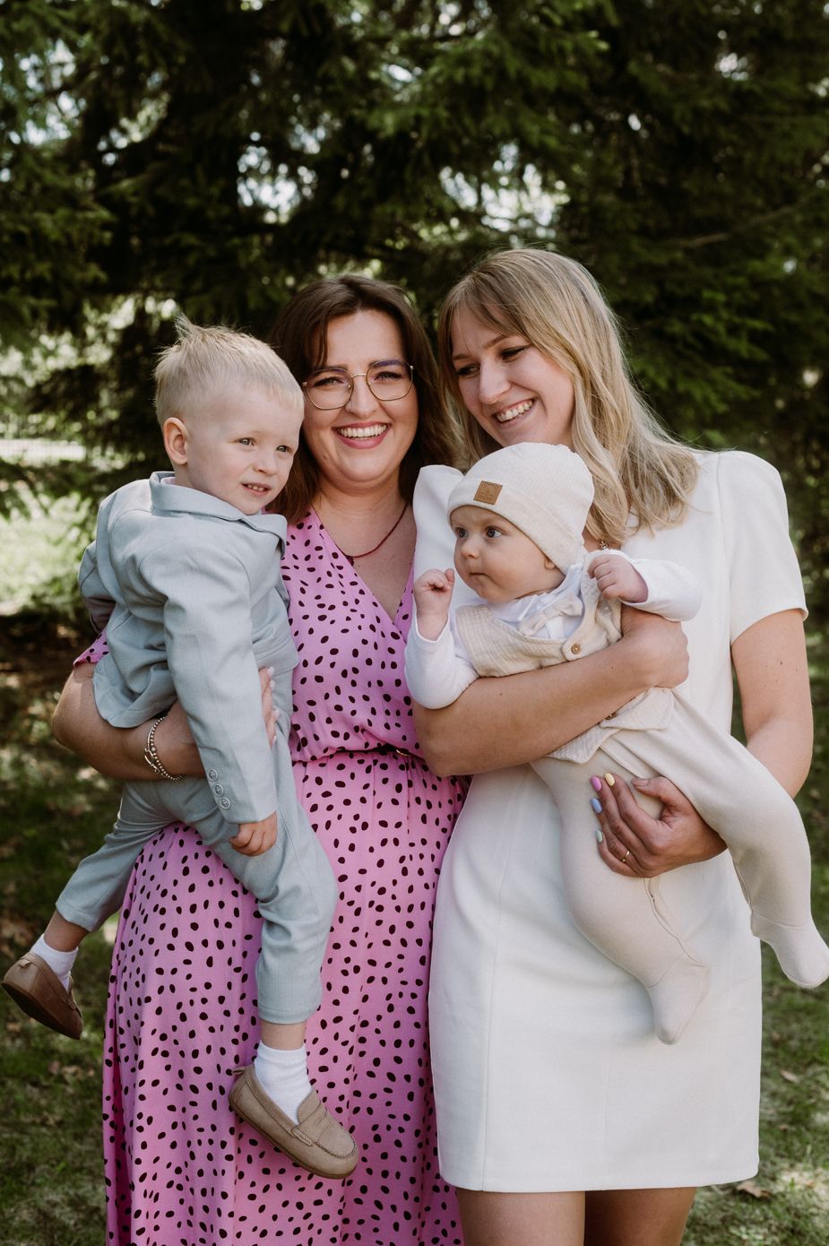 Two women smiling outdoors holding babies, wearing pink and white dresses, with evergreen trees in background.