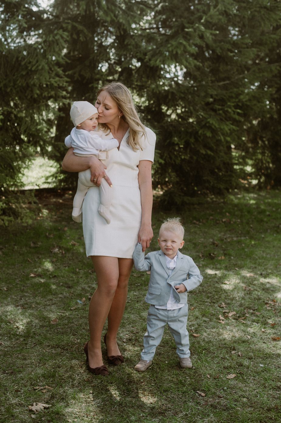 A woman in a white dress holds a baby while a toddler in a gray suit stands beside her outdoors.