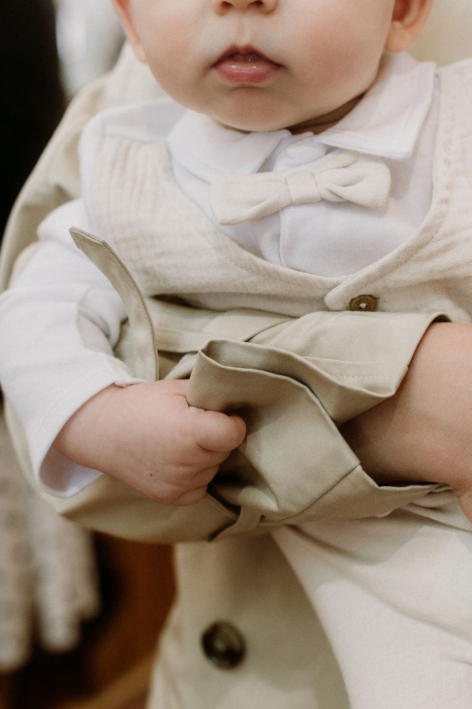 Adorable baby in a cream bow tie and vest outfit, holding fabric with chubby hands at a formal event.