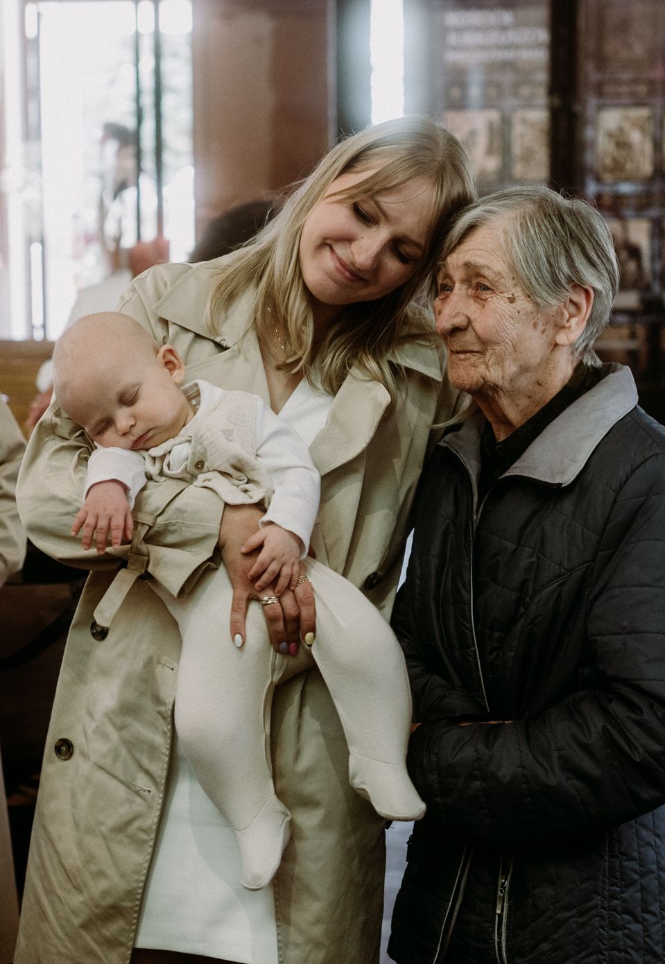 Young woman holding a baby in white outfit stands beside an elderly woman in a warm indoor setting.