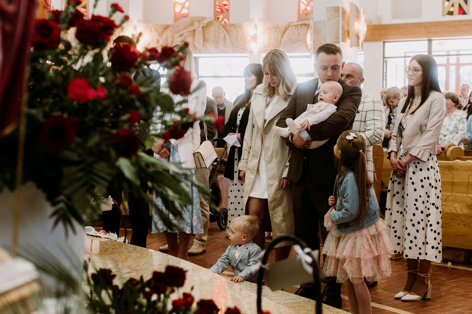 Families gathered inside a church decorated with red roses during a baptism ceremony.