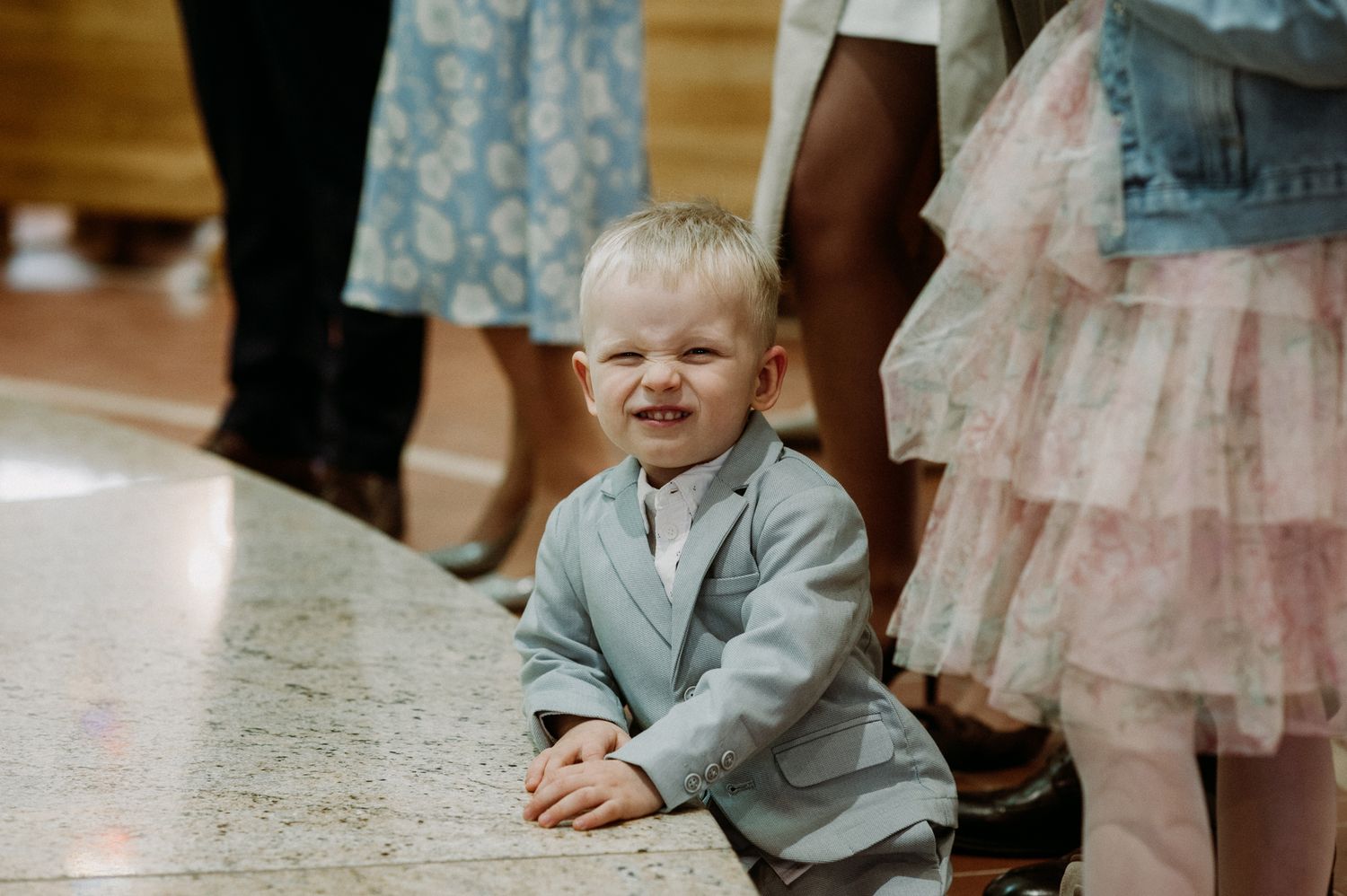 Adorable toddler boy in a gray suit sitting on the floor smiling at a wedding ceremony.