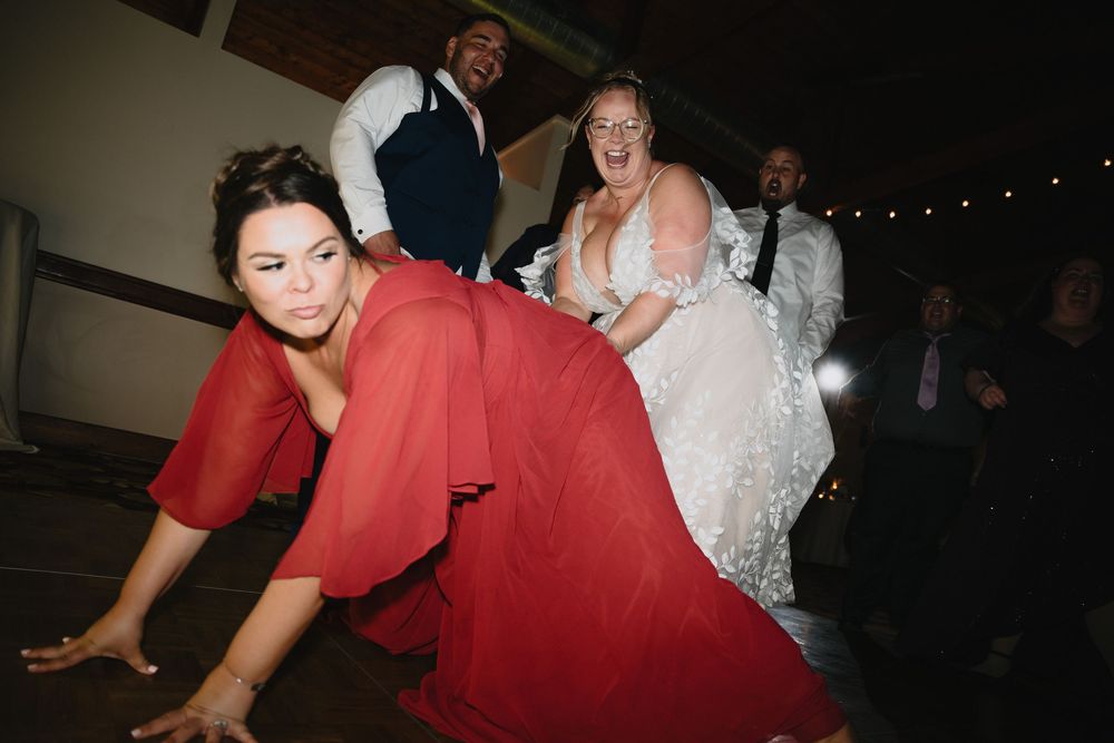 Bride in white gown laughs with guests as woman in red jumpsuit dances on the floor at a lively wedding reception.