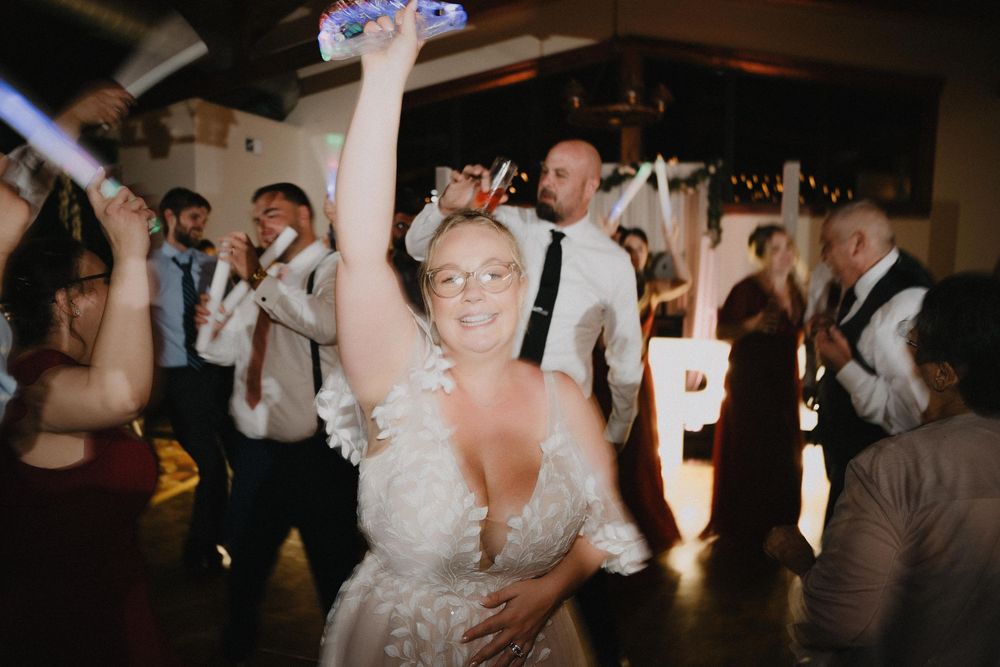 Bride in white gown dancing joyfully with raised arm at wedding reception surrounded by guests on dance floor.