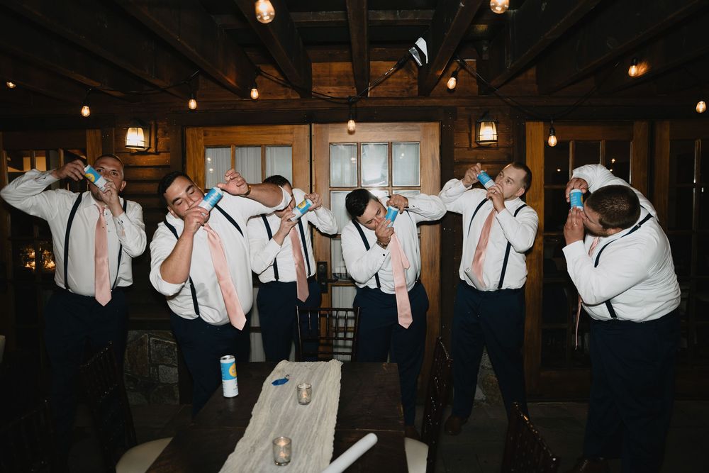 Groomsmen in white shirts and suspenders doing shots together at a rustic wedding venue celebration.