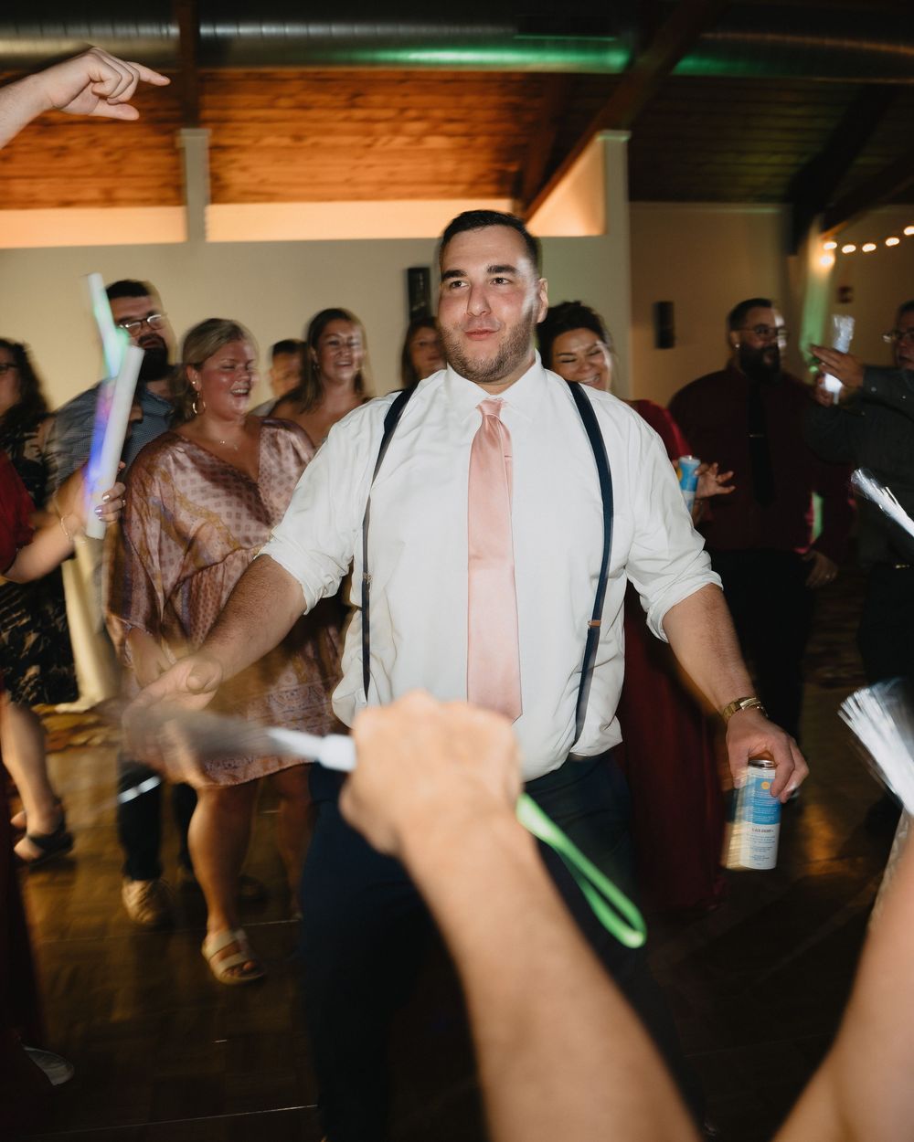 Groom in white shirt with suspenders and pink tie celebrates on wedding dance floor with guests holding glow sticks.