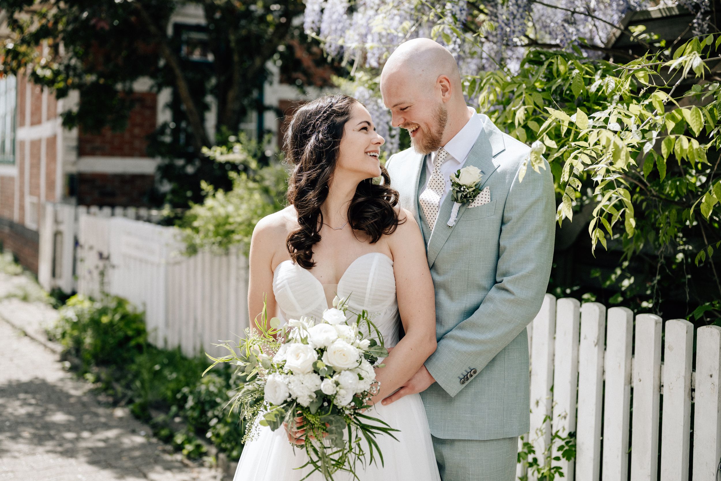 Bride and groom share a romantic moment outdoors, she holds white bouquet, he wears sage suit, white picket fence behind them.
