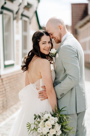 Smiling bride in white strapless gown holds white bouquet as groom in light gray suit embraces her outdoors.