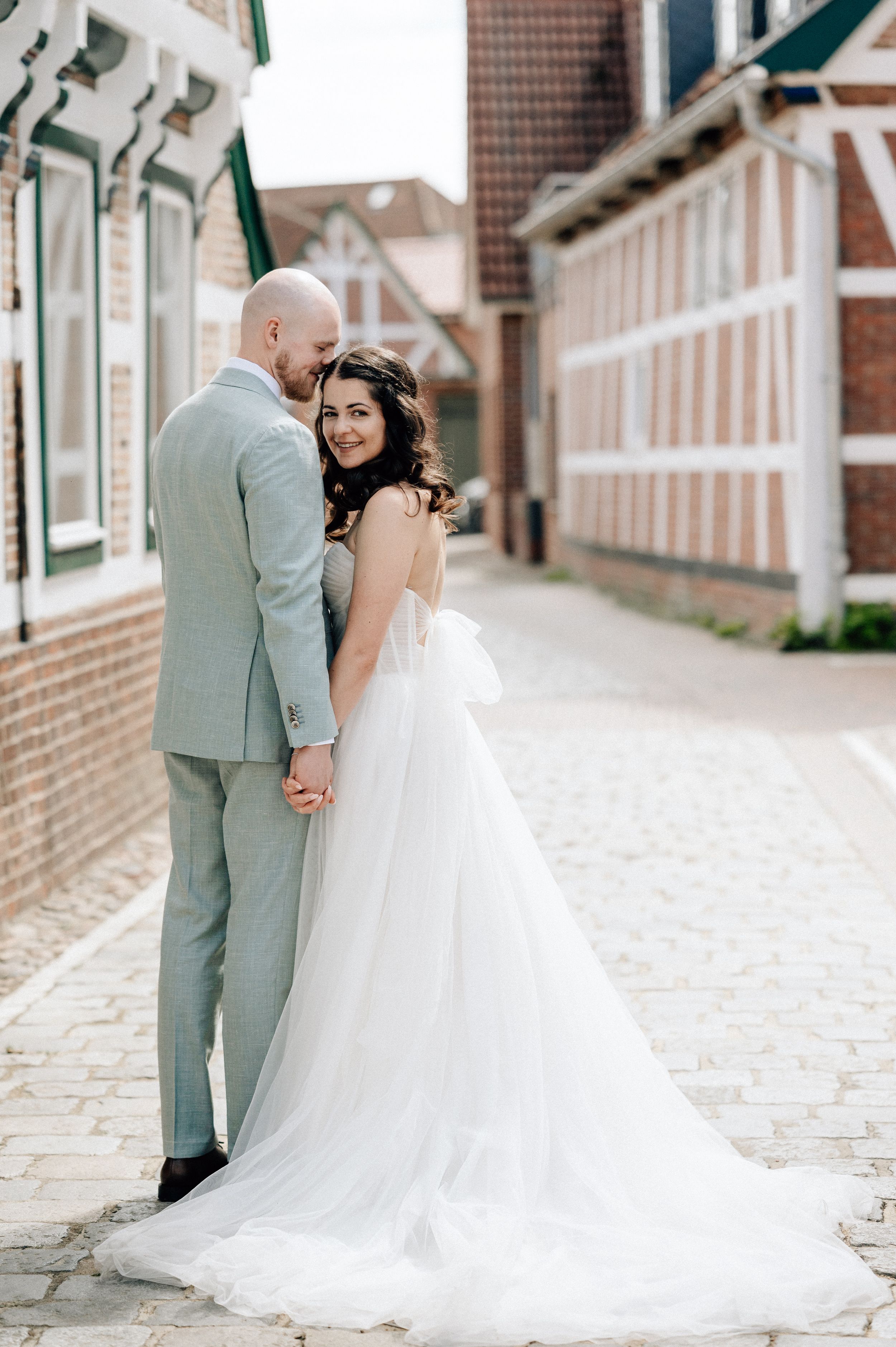 Bride in flowing white tulle gown holds hands with groom in gray suit on cobblestone street.