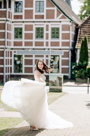 Bride in flowing white wedding dress twirling outdoors in front of a brick building on a sunny day.