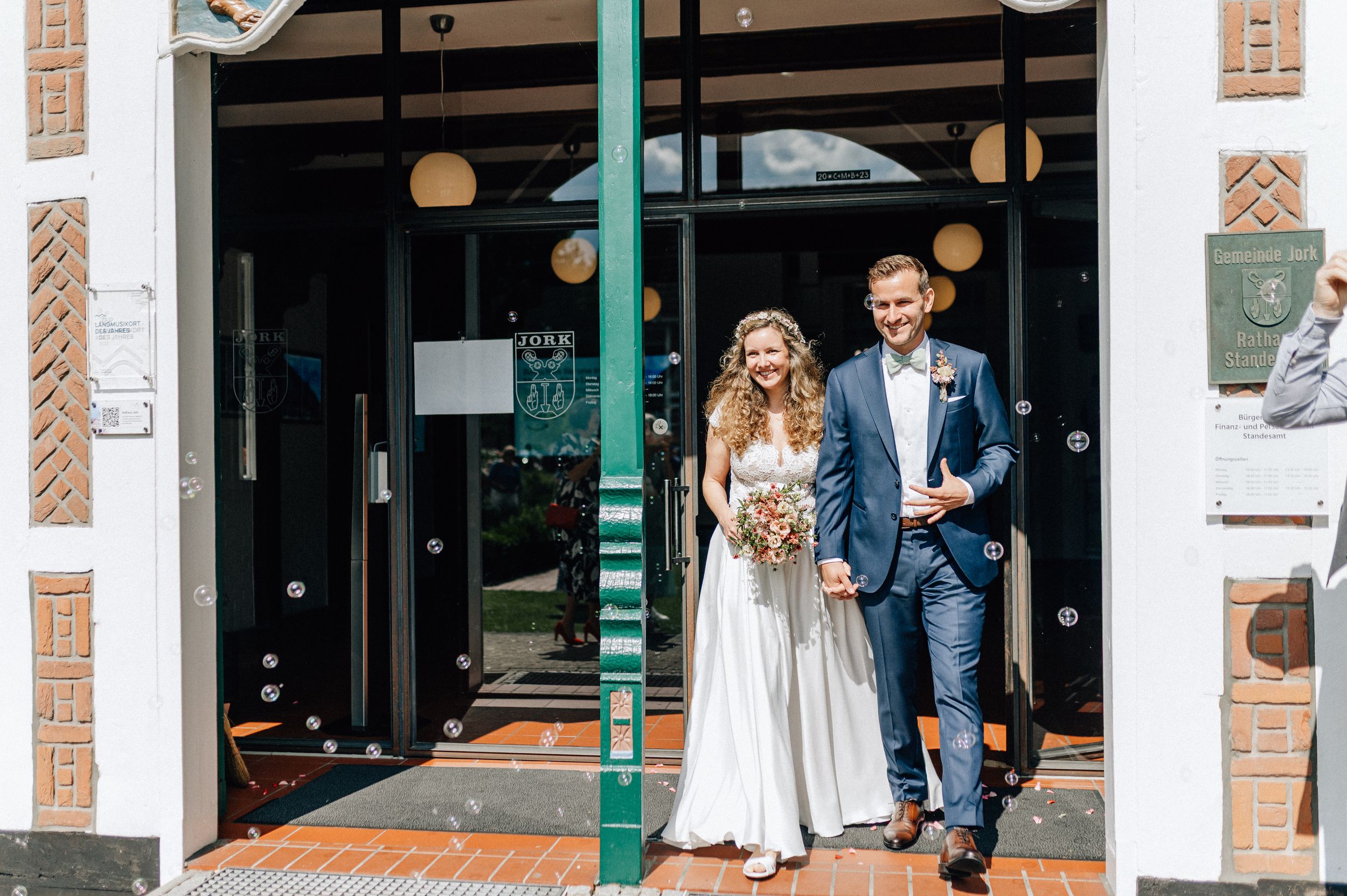 Happy newlywed couple exiting a wedding venue, bride in white gown holding bouquet, groom in blue suit.