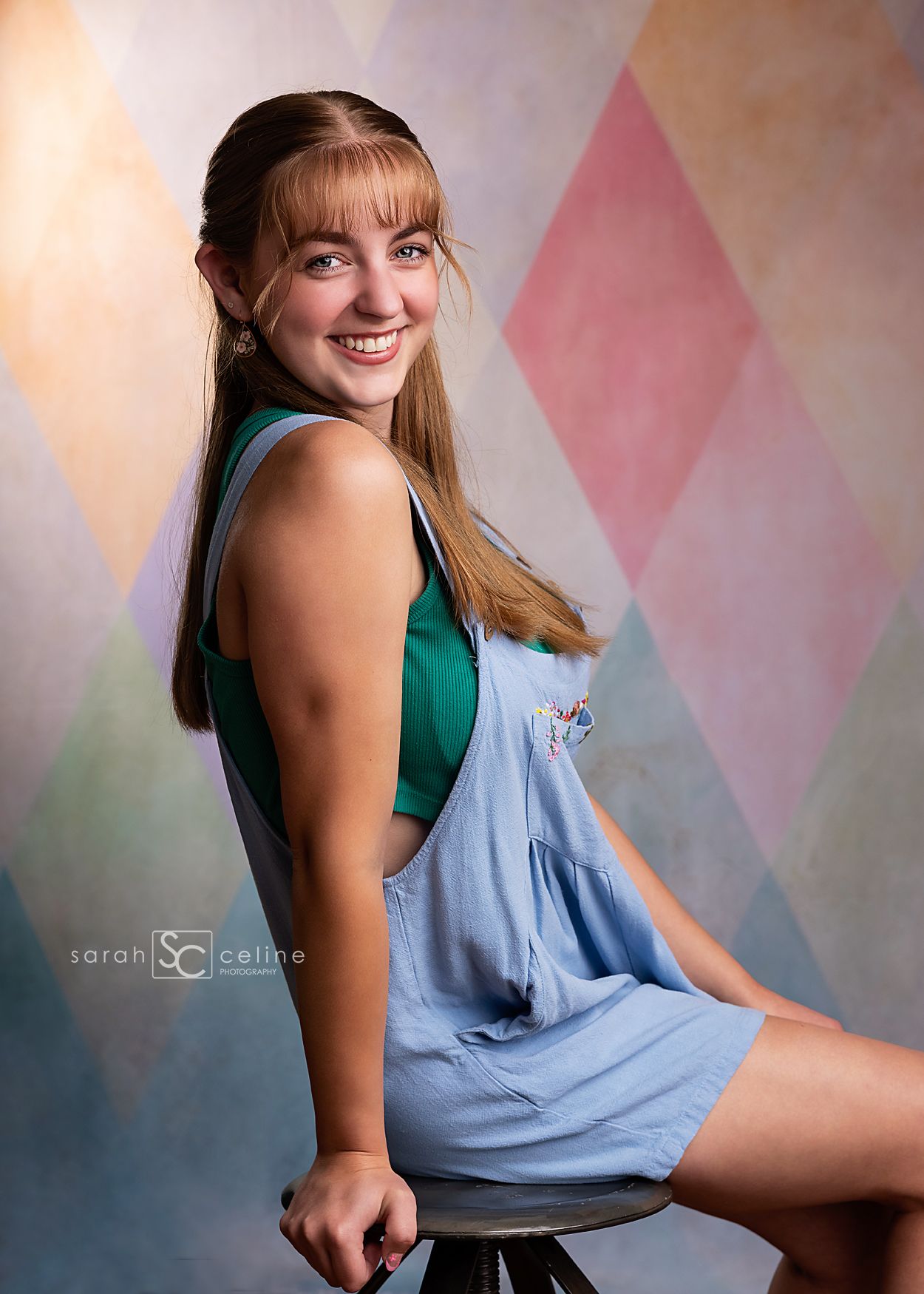 Teen girl in blue overalls and green top smiles over shoulder, seated on stool before colorful diamond pattern backdrop.