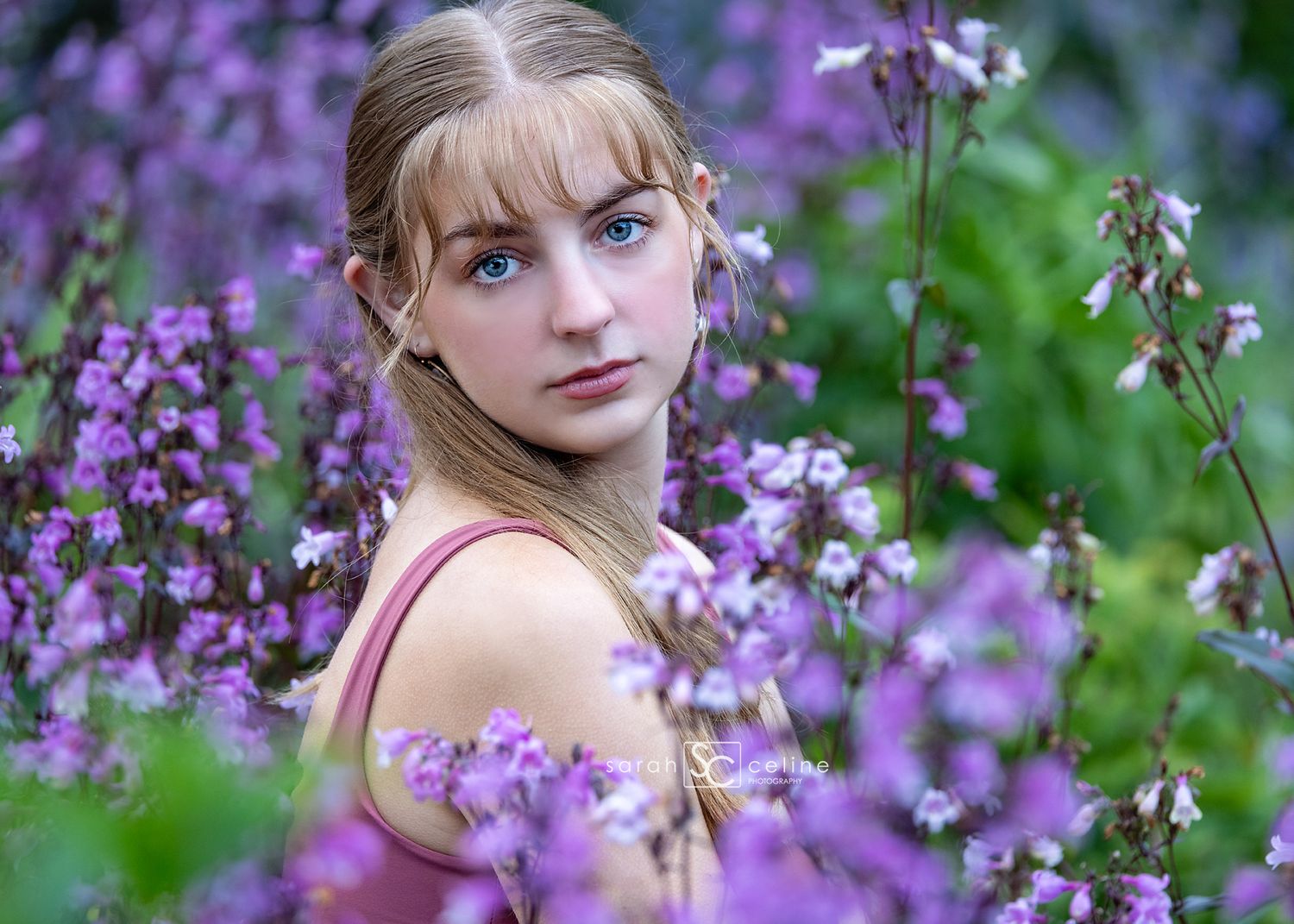 Young woman with blue eyes surrounded by purple wildflowers in a lush garden setting.