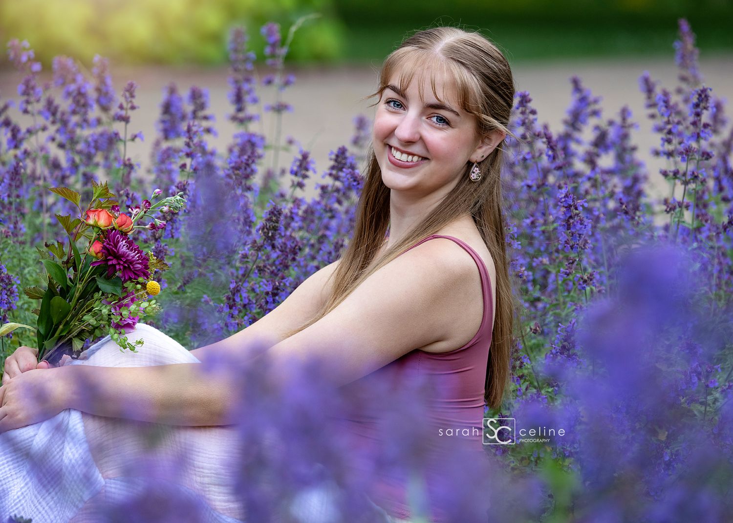 Smiling young woman sitting in a vibrant purple lavender field holding a colorful bouquet of flowers.